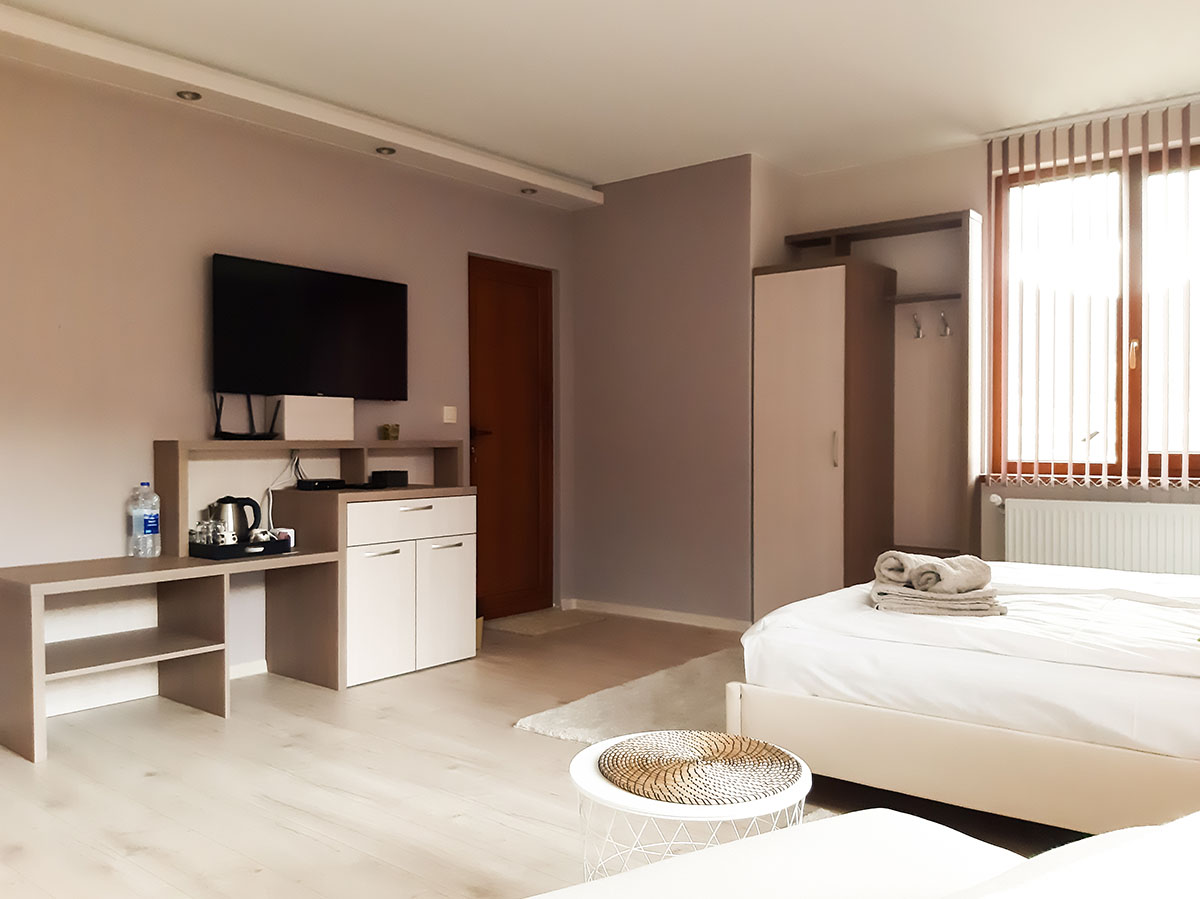 Lyst og hyggeligt hotelværelse med to senge, tv på væggen, lille køkkenområde med elkedel og garderobeskab.